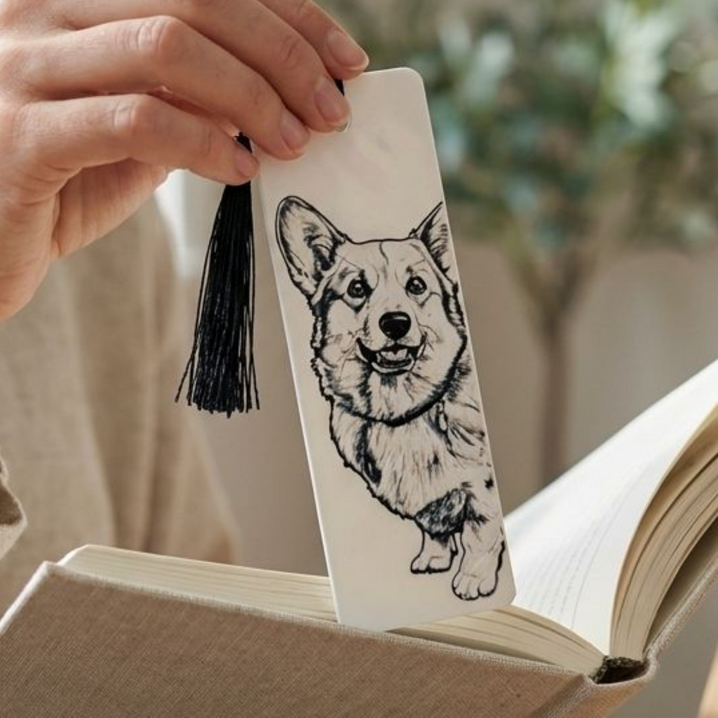 A hand holds a white bookmark featuring a black line art illustration of a corgi dog, complete with a black tassel, positioned over an open book.