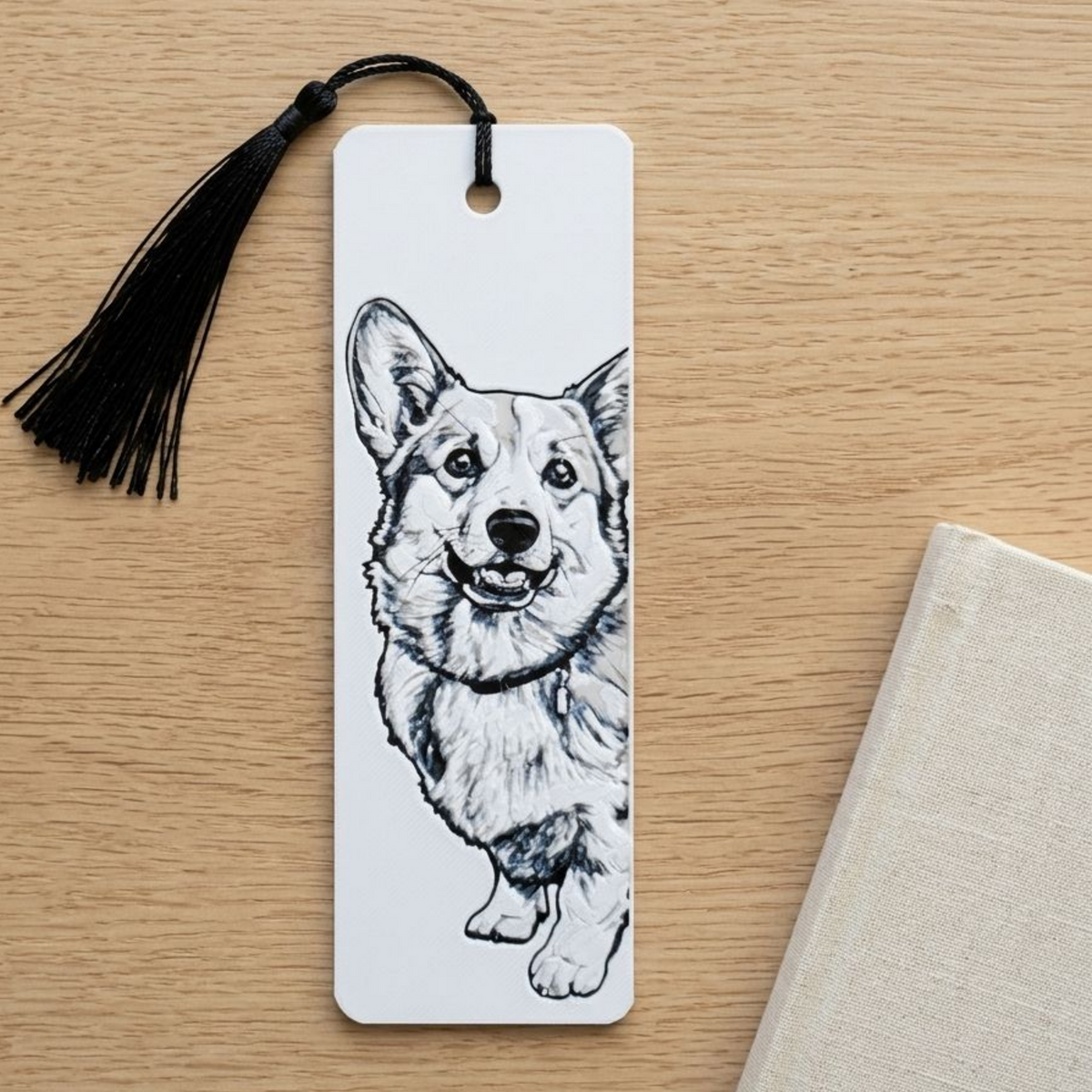A white rectangular bookmark with a black tassel hangs on a light wood tabletop. The bookmark features a black line drawing of a corgi dog.