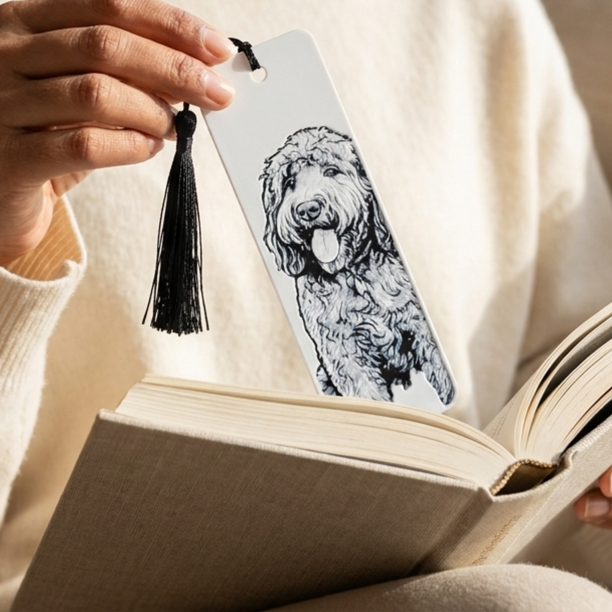 A close-up lifestyle shot of a hand holding a white bookmark with a black tassel and a black-and-white sketch of a curly-haired dog, positioned over an open book.