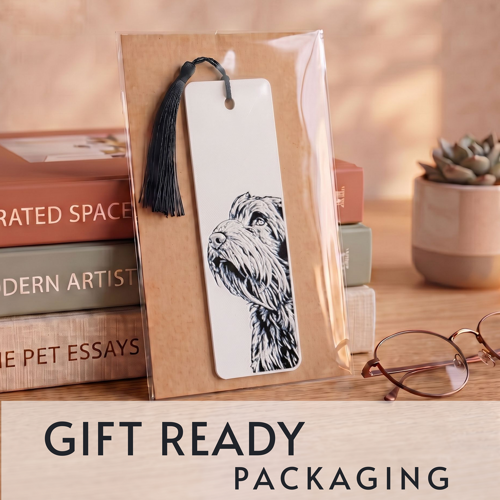 A white bookmark with a black line drawing of a dog and a black tassel, shown inside a clear gift wrap package on a wooden desk. Books, eyeglasses, and a small succulent are in the background, with the text 'GIFT READY PACKAGING' at the bottom.