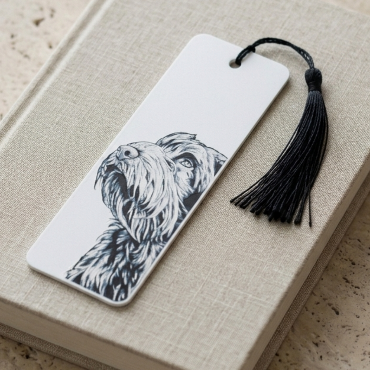 A white rectangular bookmark with a black ink illustration of a shaggy dog, resting on a beige textured book cover and attached to a black tassel.