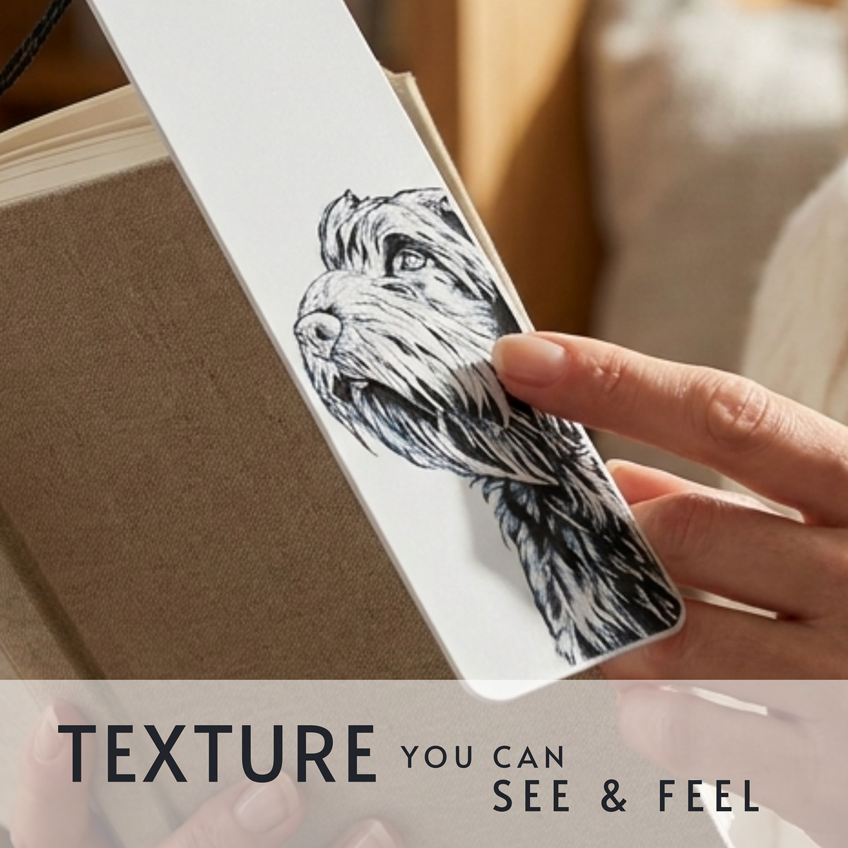 A close-up of a hand holding a white bookmark or card featuring a detailed black-and-white sketch of a dog, with a brown notebook or book cover behind it. Text at the bottom reads 'TEXTURE YOU CAN SEE & FEEL'.