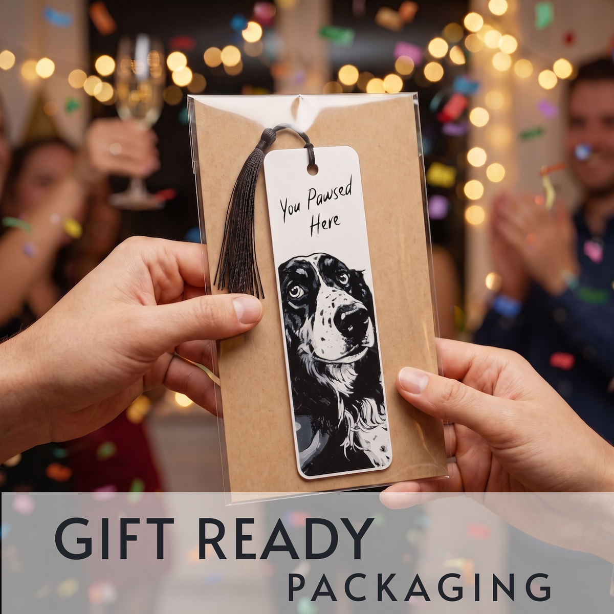 A gift-ready bookmark in kraft paper packaging with a black tassel, held by two hands. The bookmark has a white label featuring a black-and-white dog illustration and the text 'You Pawsed Here'.