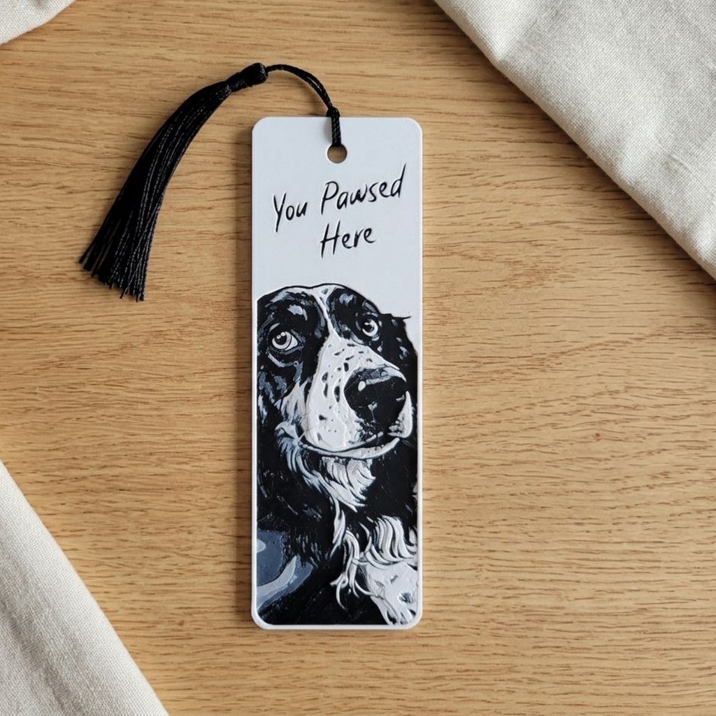 A custom rectangular bookmark featuring a black and white illustration of a dog and the handwritten text 'You Pawsed Here', complete with a black tassel, displayed on a light wood tabletop with neutral fabric props.