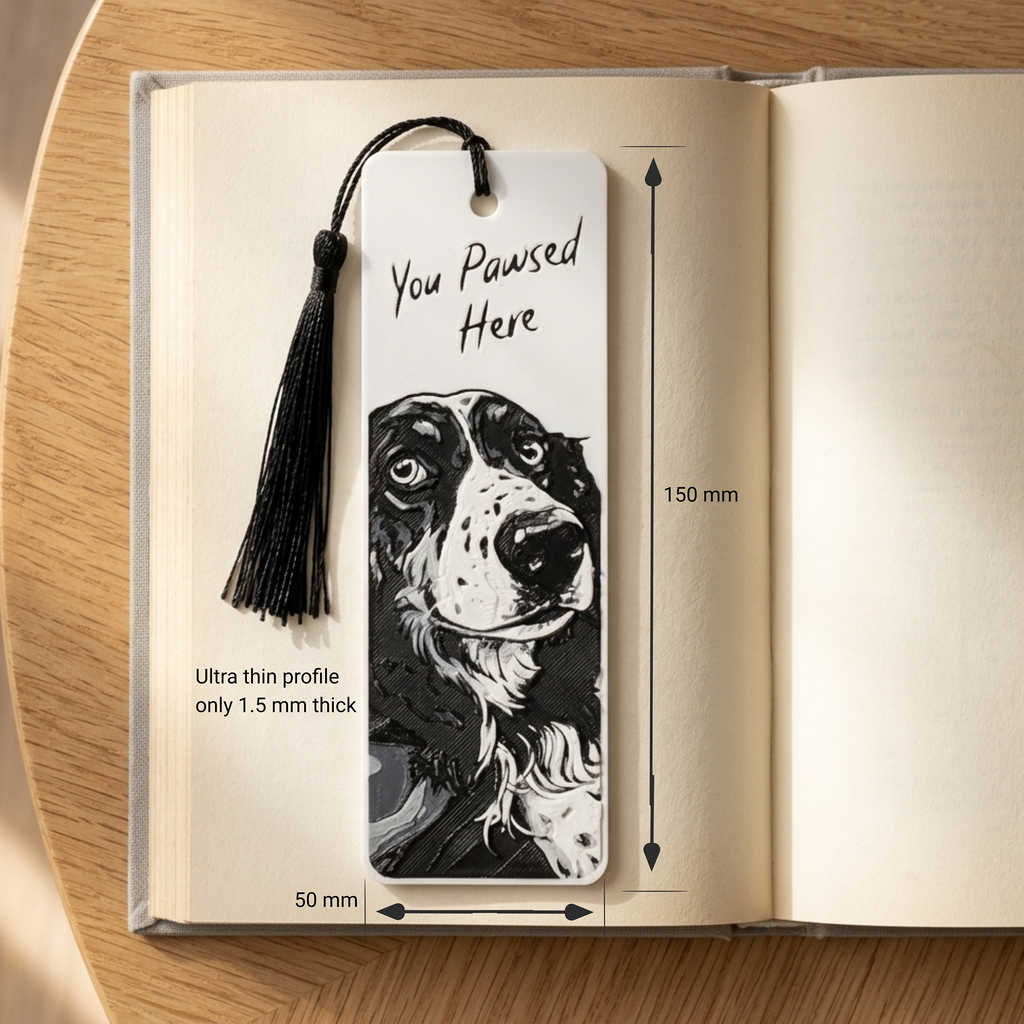 A slim white bookmark with a black tassel and a black-and-white dog portrait printed on it, shown resting on an open book. The image includes dimension arrows and text noting 150 mm length, 50 mm width, and a thin 1.