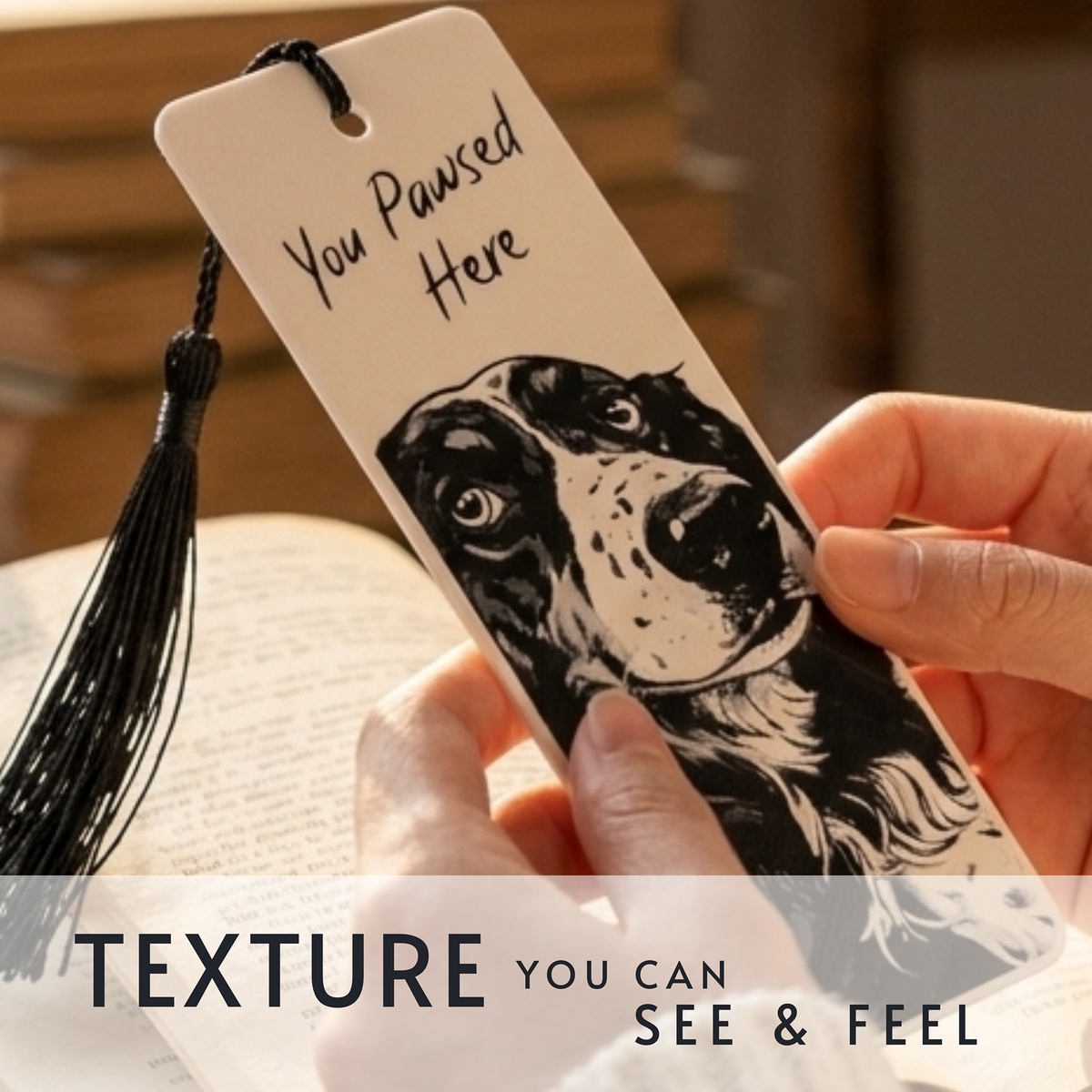 A hand holds a cream-colored bookmark featuring a black-and-white dog illustration and the text 'You Pawsed Here,' with a black tassel. The scene includes a book and warm indoor background, with overlaid text reading 'TEXTURE YOU CAN SEE & FEEL.