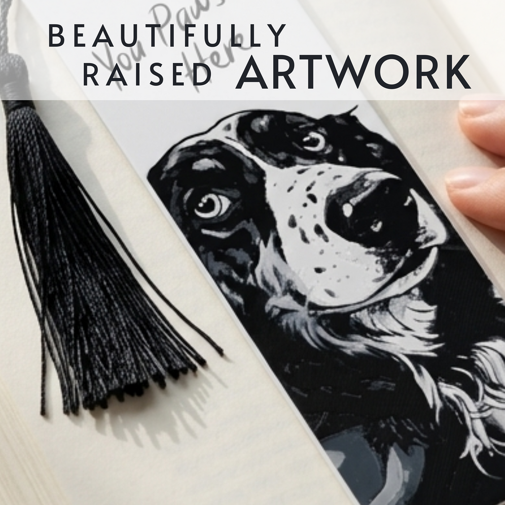 A close-up of a black-and-white printed bookmark featuring a dog portrait, placed on cream-colored pages with a black tassel bookmark nearby. A hand is visible at the right edge, and overlaid text reads 'BEAUTIFULLY RAISED ARTWORK'.