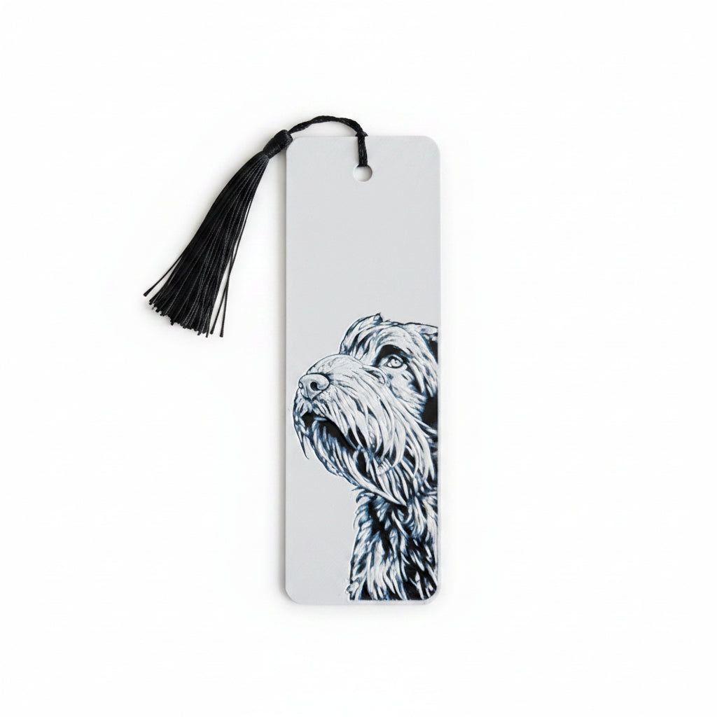 A white rectangular 3D printed bookmark with a black tassel at the top and a black-and-white illustration of a handsome scruffy dog on the right side, photographed against a light beige textured background.